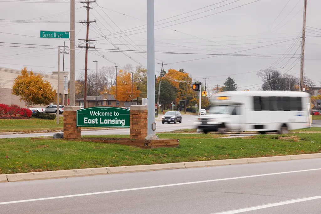 A bus travels past the East Lansing sign on Grand River Avenue, where Michigan Auto Law helps crash victims as a bus accident lawyer in East Lansing.