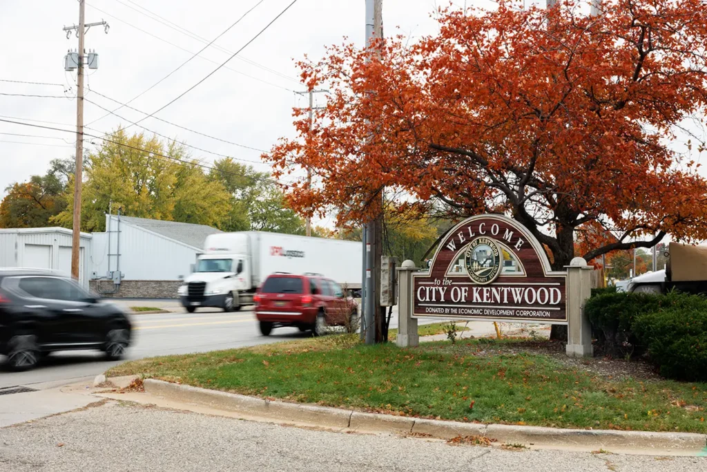 Traffic moves past the Kentwood welcome sign beneath fall trees, representing the area served by a car accident attorney in Kentwood.