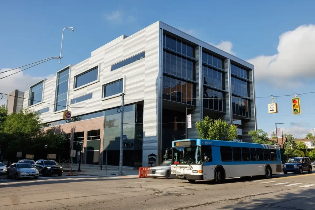 Modern office building with a city bus traveling along a busy Ann Arbor street, highlighting Michigan Auto Law truck accident lawyer services and roadways similar to those the firm handles.